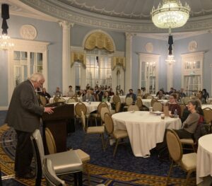 Michigan_Kevin_Speech NCSA Members gather in a blue ball room. Individuals sit at round tables across the room. A speaker stands at a podium in the foreground.